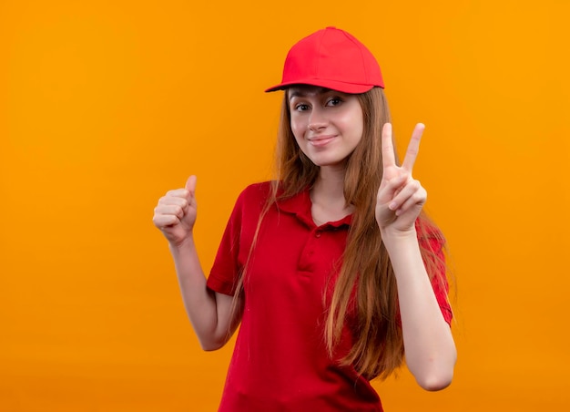 Jovem entregadora de uniforme vermelho satisfeita fazendo o sinal da paz e exibindo o polegar em um espaço laranja isolado com espaço de cópia