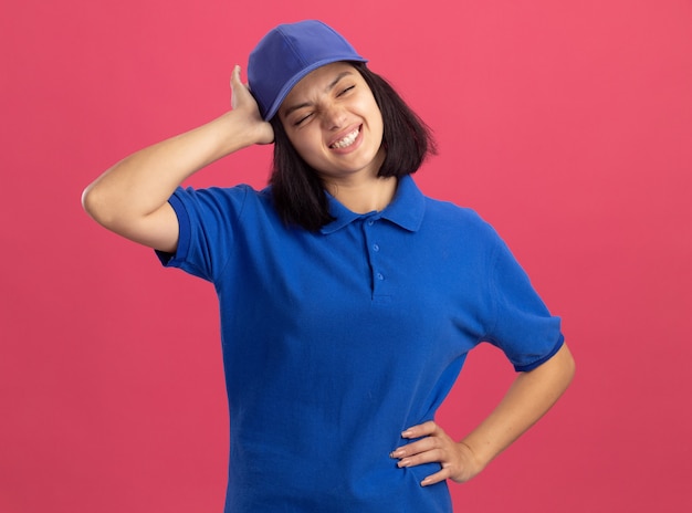 Foto grátis jovem entregadora descontente com uniforme azul e boné tocando a cabeça, com dor de cabeça em pé sobre a parede rosa