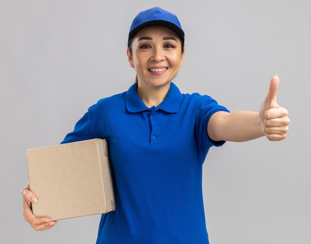 Jovem entregadora feliz com uniforme azul e boné segurando uma caixa de papelão com um sorriso no rosto mostrando os polegares para cima