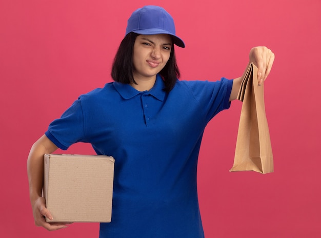 Jovem entregadora insatisfeita com uniforme azul e boné segurando um pacote de papel e uma caixa de papelão, parecendo fazer uma boca irônica em pé sobre a parede rosa