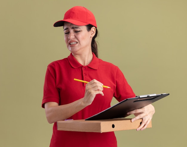 Jovem entregadora irritada de uniforme e boné segurando uma prancheta de pacote de pizza e um lápis olhando para baixo