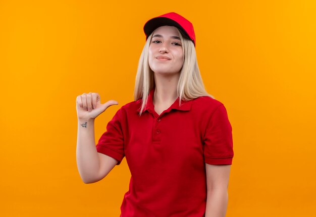 Jovem entregadora satisfeita com camiseta vermelha e boné apontando para ela mesma em um fundo laranja isolado