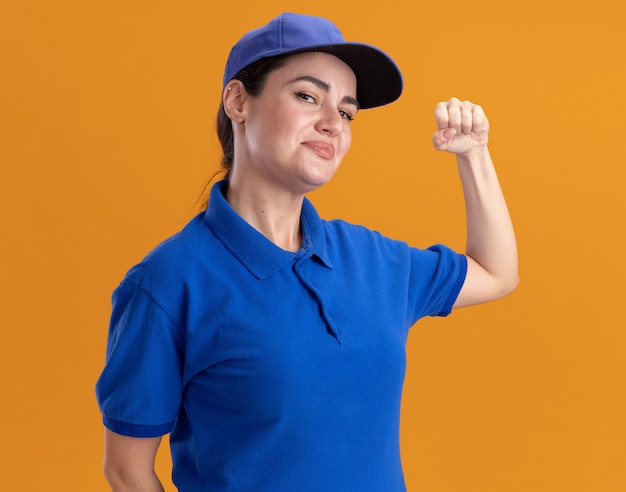 Jovem entregadora satisfeita de uniforme e boné, fazendo gesto de bater na porta, isolada na parede laranja