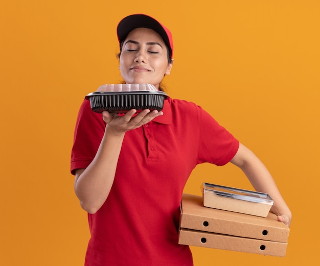 Jovem entregadora satisfeita de uniforme e boné, segurando caixas de pizza e cheirando recipientes de comida na mão, isolada na parede laranja