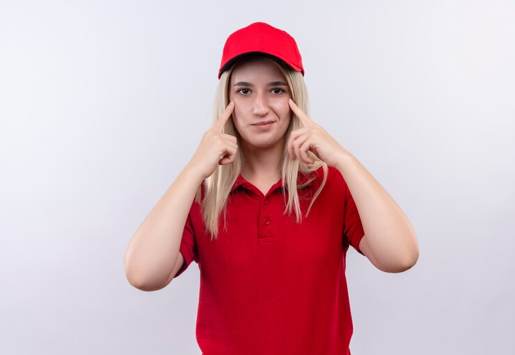 Foto grátis jovem entregadora satisfeita, vestindo camiseta vermelha e boné, colocando os dedos nos olhos em um fundo branco isolado
