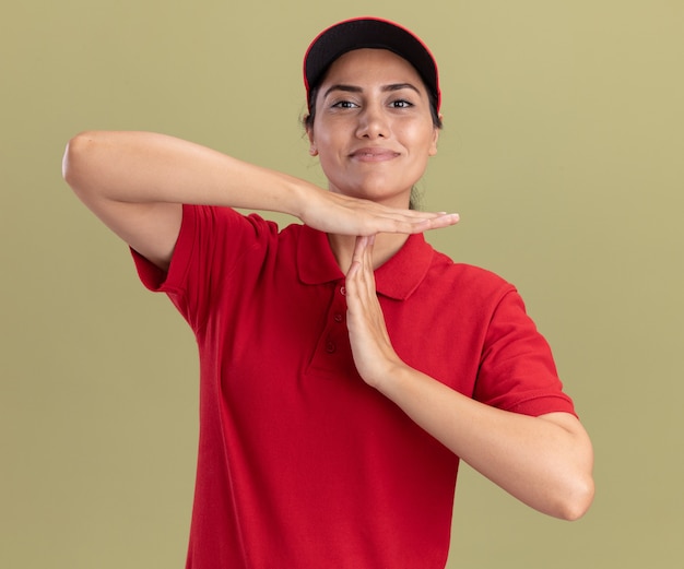 Jovem entregadora satisfeita vestindo uniforme com tampa mostrando gesto de tempo limite isolado na parede verde oliva