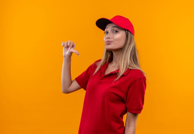 Jovem entregadora satisfeita vestindo uniforme vermelho e boné apontando para si mesma