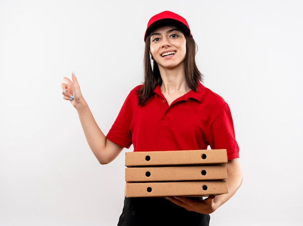 Jovem entregadora satisfeita vestindo uniforme vermelho e boné segurando uma pilha de caixas de pizza apontando com o dedo indicador para o lado sorrindo alegremente em pé sobre um fundo branco