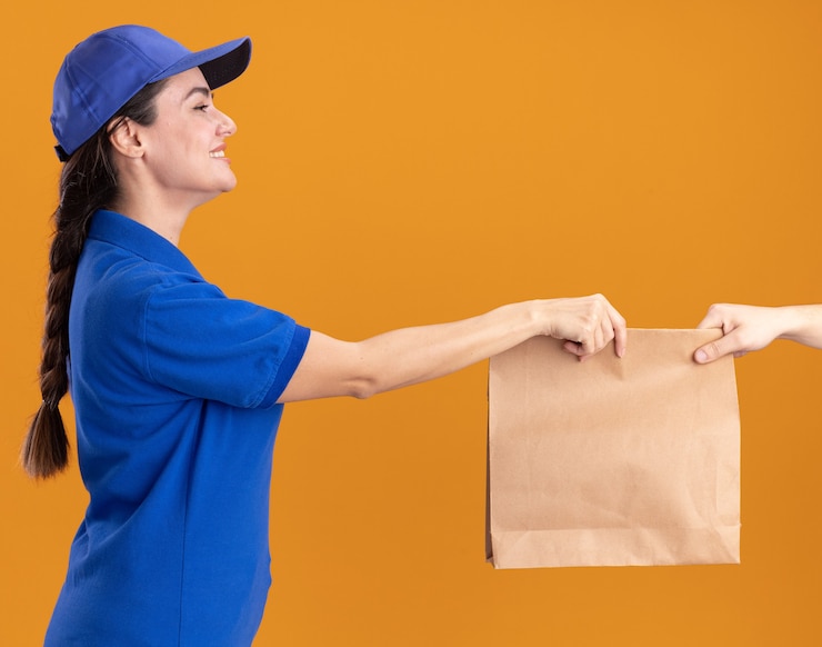 Foto grátis jovem entregadora sorridente de uniforme e boné em vista de perfil, dando um pacote de papel para o cliente, olhando para o cliente