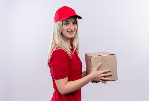 Jovem entregadora sorridente usando camiseta vermelha e boné no aparelho dentário segurando uma caixa no fundo branco isolado