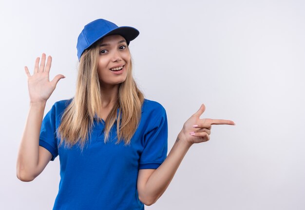 Jovem entregadora sorridente usando uniforme azul e boné mostrando gesto de parada e pontos para o lado isolado na parede branca com espaço de cópia