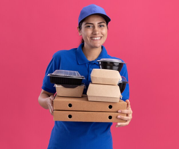 Jovem entregadora sorridente, usando uniforme com tampa, segurando recipientes de comida em caixas de pizza isoladas na parede rosa