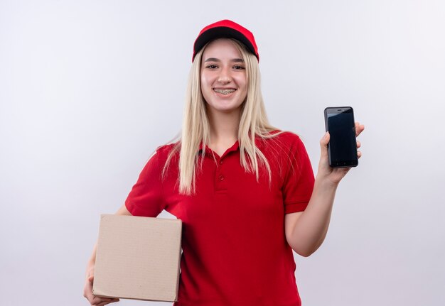 Jovem entregadora sorridente, vestindo camiseta vermelha e boné com aparelho dentário segurando o telefone e a caixa no fundo branco isolado