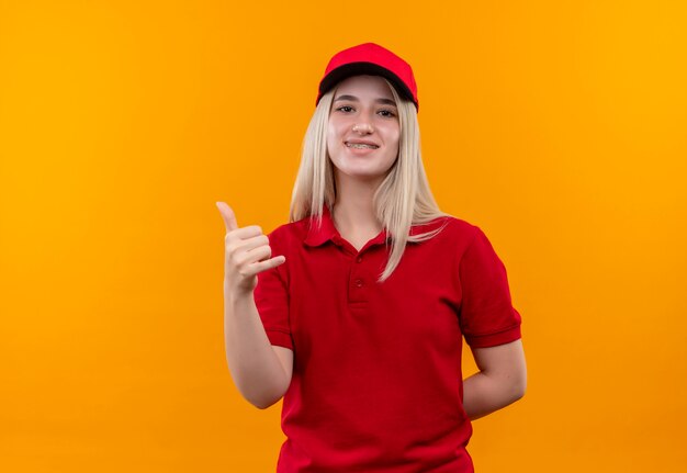 Jovem entregadora sorridente, vestindo camiseta vermelha e boné no aparelho dentário, mostrando gesto de telefone em fundo laranja isolado