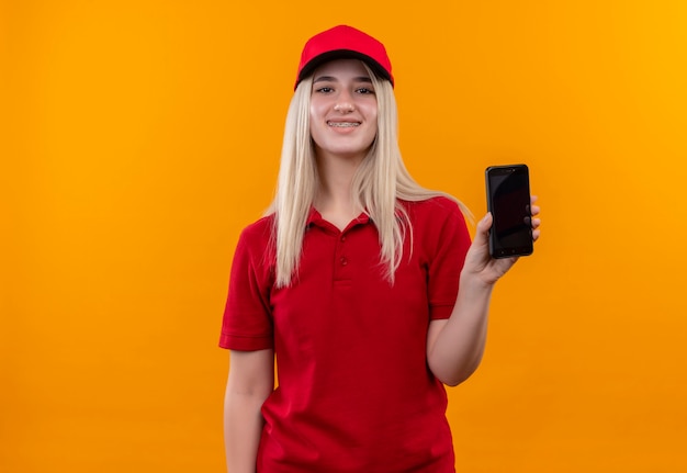 Jovem entregadora sorridente, vestindo camiseta vermelha e boné no aparelho dentário, mostrando o telefone para a câmera em fundo laranja isolado