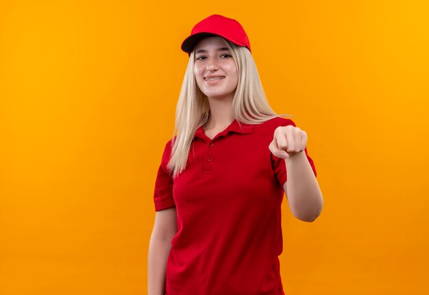 Jovem entregadora sorridente, vestindo camiseta vermelha e boné, segurando o punho para a câmera em um fundo laranja isolado