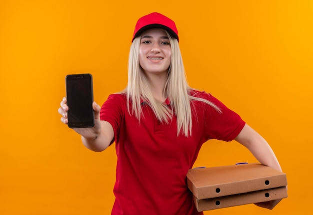 Jovem entregadora sorridente, vestindo camiseta vermelha e boné, segurando uma caixa de pizza e mostrando o telefone para a câmera em fundo laranja isolado