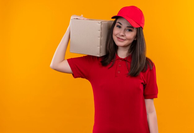 Jovem entregadora sorridente, vestindo uma camiseta vermelha com tampa vermelha na caixa de ombro na parede laranja isolada