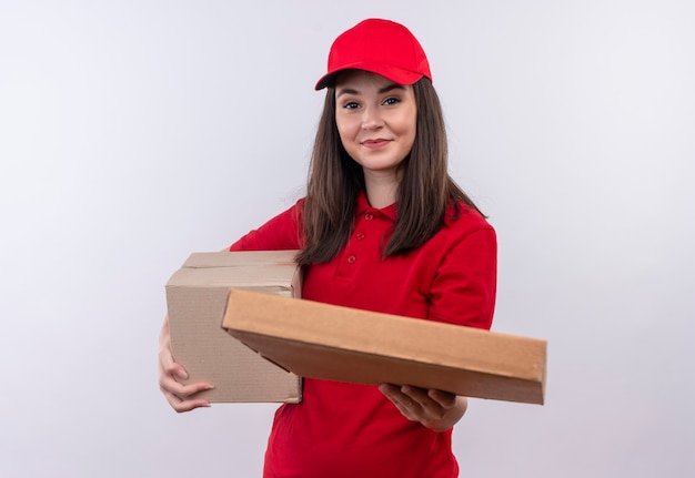 Jovem entregadora sorridente, vestindo uma camiseta vermelha com tampa vermelha, segurando uma caixa e segurando a caixa de pizza na parede branca isolada