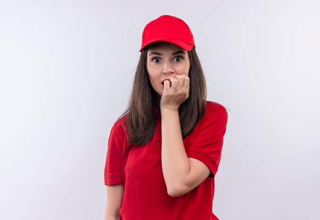Jovem entregadora surpresa, vestindo uma camiseta vermelha com boné vermelho, agarrou o queixo na parede branca isolada