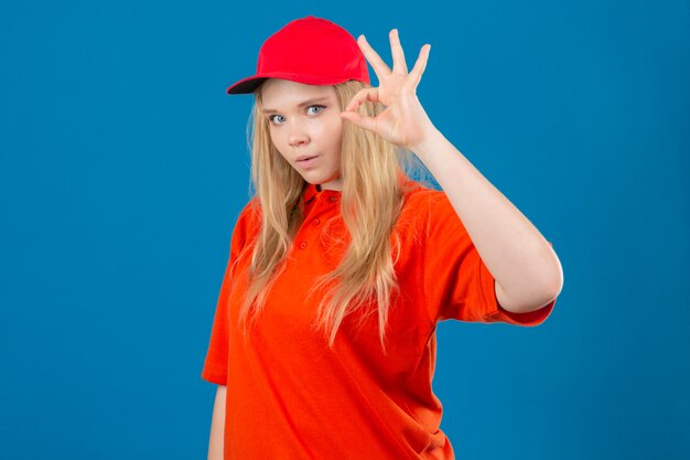 Jovem entregadora vestindo camisa pólo laranja e boné vermelho, parecendo surpresa fazendo sinal de ok sobre fundo azul isolado