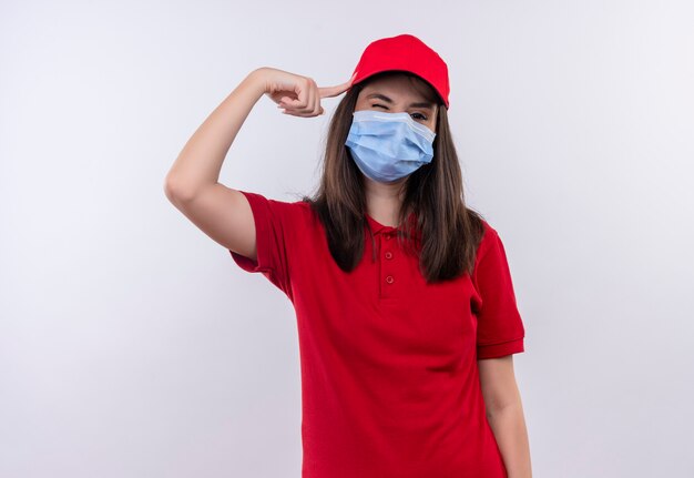 Jovem entregadora vestindo camiseta vermelha com boné vermelho e máscara facial colocou o dedo na cabeça sobre fundo branco isolado