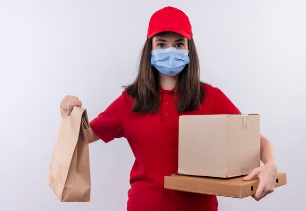 Jovem entregadora vestindo camiseta vermelha com boné vermelho e máscara facial segurando um pacote, uma caixa e uma caixa de pizza em um fundo branco isolado