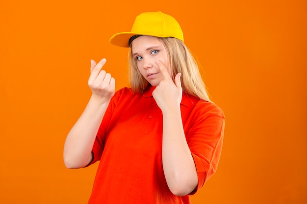 Jovem entregadora vestindo uma camisa polo vermelha e boné amarelo fazendo gesto de dinheiro sorrindo sobre fundo laranja isolado