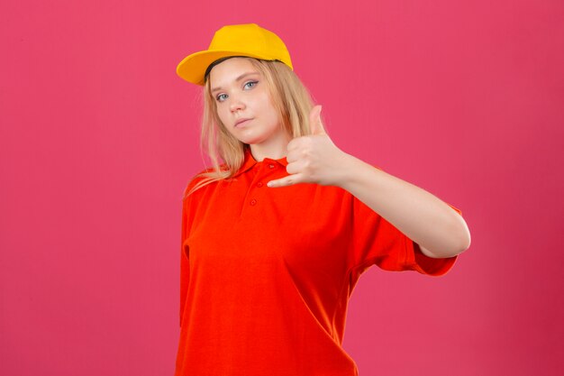Jovem entregadora vestindo uma camisa pólo vermelha e boné amarelo, fazendo um gesto de me ligar parecendo confiante sobre um fundo rosa isolado jpg