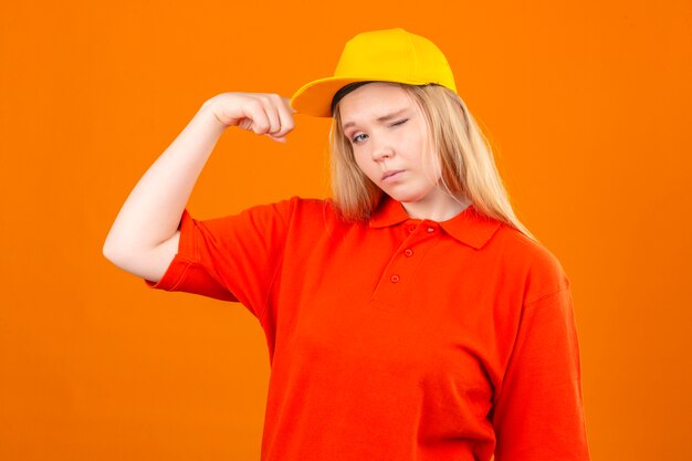 Jovem entregadora vestindo uma camisa pólo vermelha e boné amarelo piscando em pé com o conceito de vencedor de punho erguido sobre fundo laranja isolado