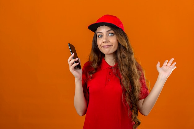 Jovem entregadora vestindo uma camisa pólo vermelha e boné segurando um celular com uma expressão confusa e sem noção com os braços e as mãos levantando o conceito de dúvida sobre um fundo laranja isolado