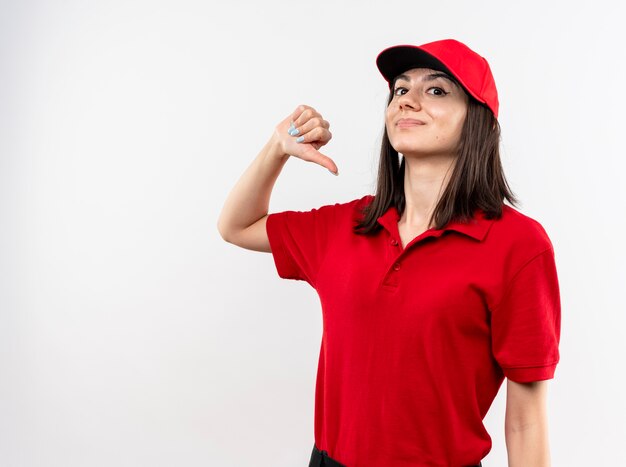 Jovem entregadora vestindo uniforme vermelho e boné apontando para si mesma, sorrindo confiante em pé sobre uma parede branca