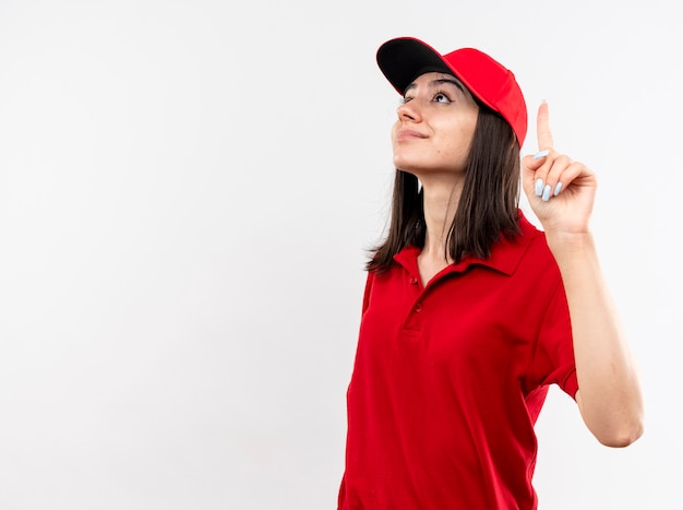 Jovem entregadora vestindo uniforme vermelho e boné olhando para cima mostrando o dedo indicador sorrindo confiante em pé sobre um fundo branco