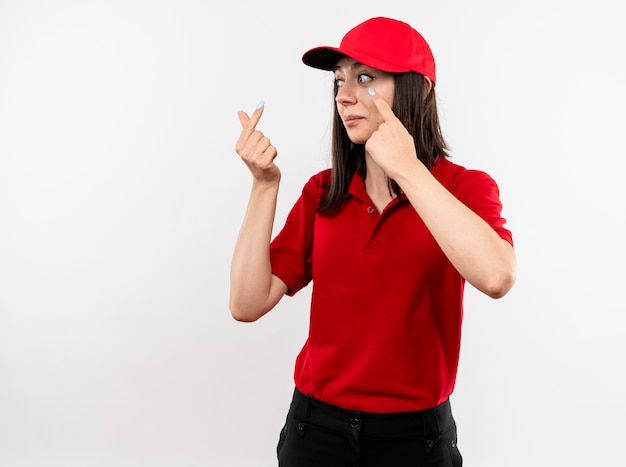 Jovem entregadora vestindo uniforme vermelho e boné olhando para o lado fazendo gesto de dinheiro esperando o pagamento em pé sobre fundo branco