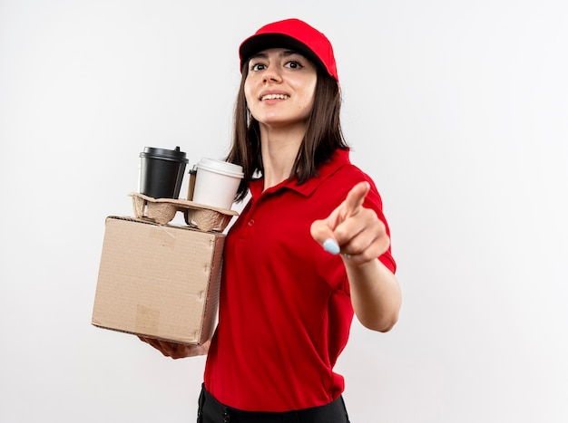 Jovem entregadora vestindo uniforme vermelho e boné segurando a embalagem da caixa e xícaras de café apontando com o dedo indicador e sorrindo com um rosto feliz em pé sobre a parede branca