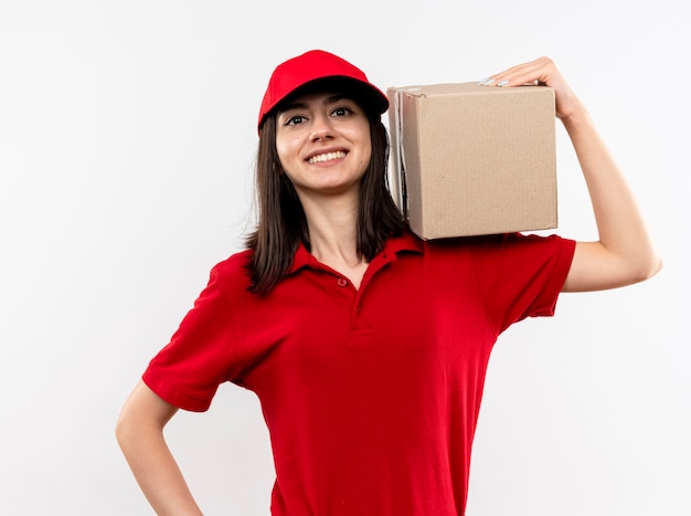 Jovem entregadora vestindo uniforme vermelho e boné segurando a embalagem da caixa, parecendo confiante com um grande sorriso no rosto, de pé sobre um fundo branco