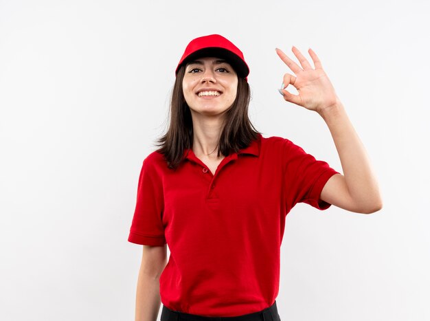 Jovem entregadora vestindo uniforme vermelho e boné sorrindo com uma cara feliz, mostrando uma placa de ok em pé sobre uma parede branca