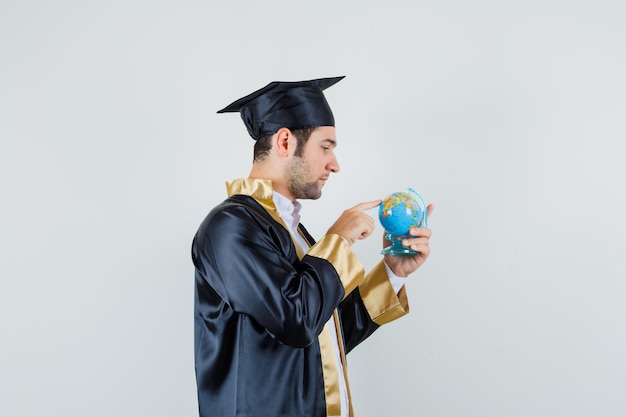 Jovem, escolhendo o destino no globo da escola em uniforme de pós-graduação e procurando focado. .