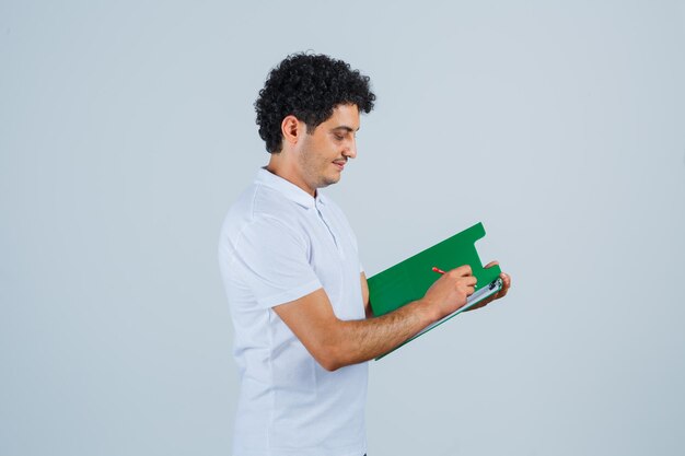 Jovem escrevendo algo no caderno em jeans e camiseta branca e olhando feliz, vista frontal.