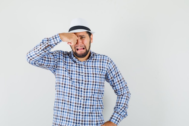 Jovem esfregando os olhos enquanto chora como uma criança de camisa xadrez, chapéu e parecendo ofendido