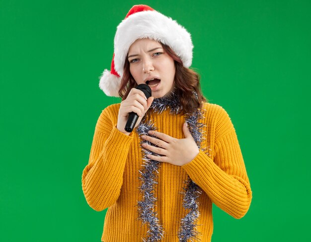 Jovem eslava confiante com chapéu de Papai Noel e guirlanda no pescoço segura o microfone fingindo cantar isolado em um fundo verde com espaço de cópia