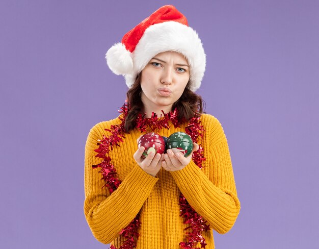 Jovem eslava decepcionada com chapéu de Papai Noel e guirlanda no pescoço segura enfeites de bola de vidro isolados em um fundo roxo com espaço de cópia