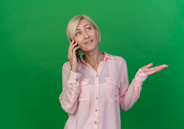 Foto grátis jovem eslava loira impressionada olhando para cima, falando no telefone e mostrando a mão vazia isolada em um fundo verde com espaço de cópia