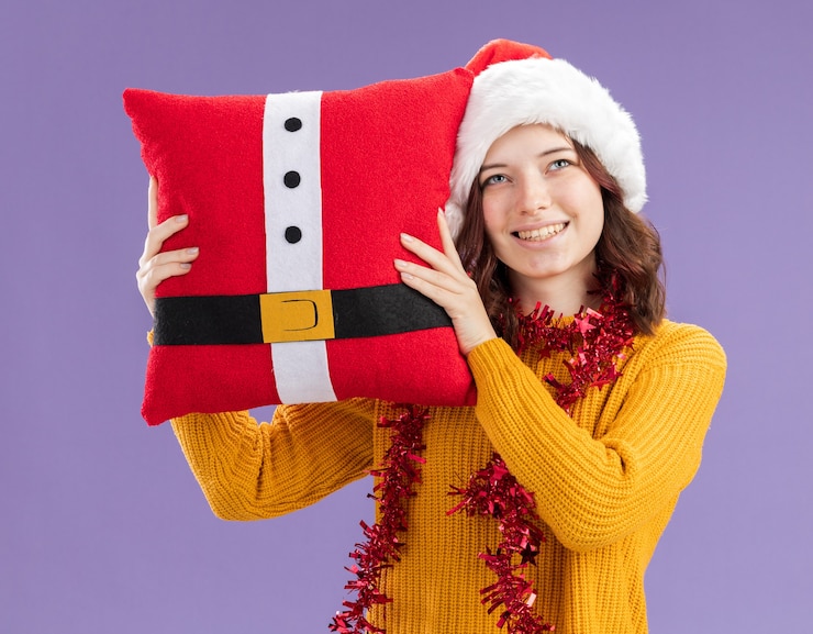 Foto grátis jovem eslava satisfeita com chapéu de papai noel e guirlanda em volta do pescoço segurando uma almofada decorada, olhando para cima isolada no fundo roxo com espaço de cópia