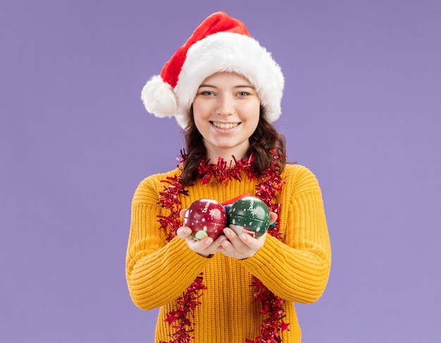 Jovem eslava sorridente com chapéu de Papai Noel e guirlanda no pescoço segurando enfeites de bola de vidro isolados na parede roxa com espaço de cópia