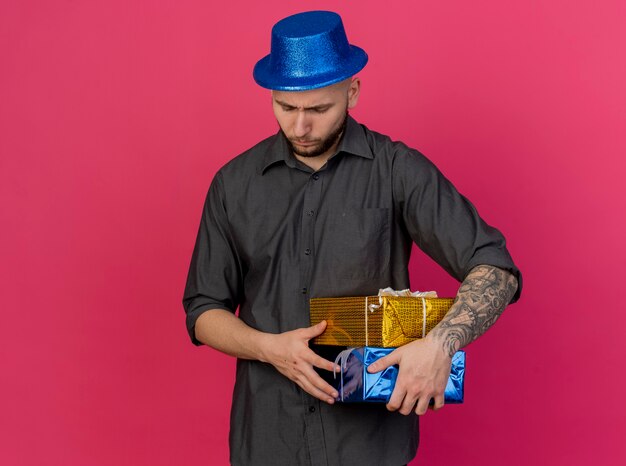 Jovem eslavo bonito carrancudo com um chapéu de festa segurando pacotes de presentes, olhando para baixo, isolados em um fundo carmesim com espaço de cópia