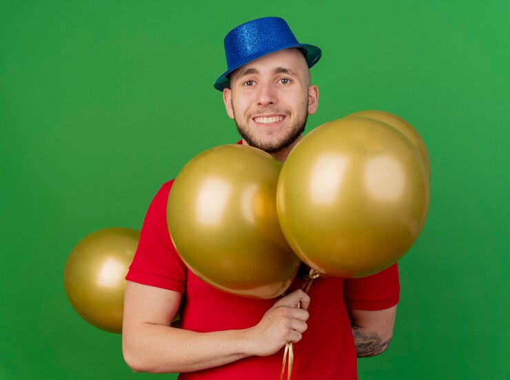 Foto grátis jovem eslavo bonito sorridente com chapéu de festa segurando balões, olhando para a câmera isolada sobre fundo verde