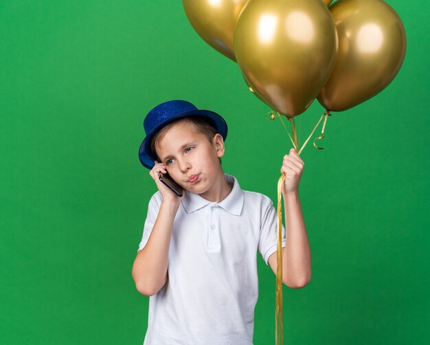 Foto grátis jovem eslavo confiante com chapéu de festa azul segurando balões de hélio e falando no telefone, olhando para o lado isolado na parede verde com espaço de cópia