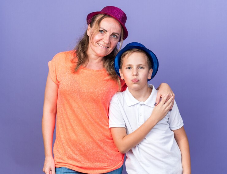 Foto grátis jovem eslavo satisfeito com chapéu de festa azul em pé com a mãe usando chapéu de festa violeta isolado na parede roxa com espaço de cópia