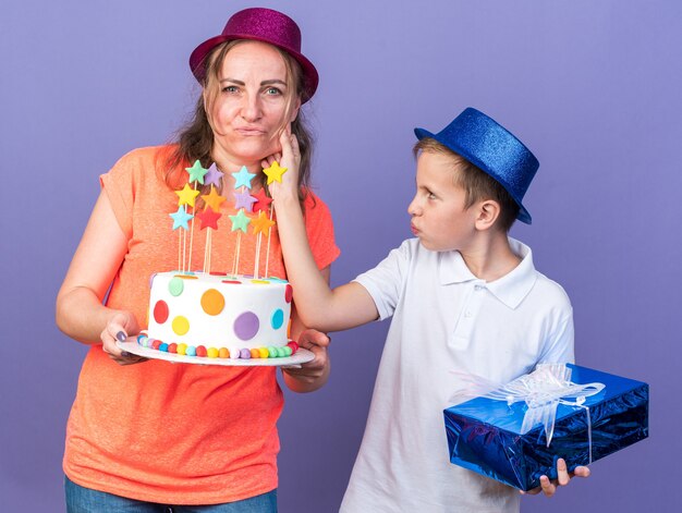 jovem eslavo satisfeito com um chapéu de festa azul segurando uma caixa de presente e puxando a bochecha de sua mãe usando um chapéu de festa violeta e segurando um bolo de aniversário isolado na parede roxa com espaço de cópia